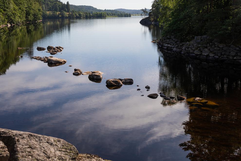 Stenar i vatten nära strandkant. Vattnet omges av grönskande växtlighet och kallmurar.