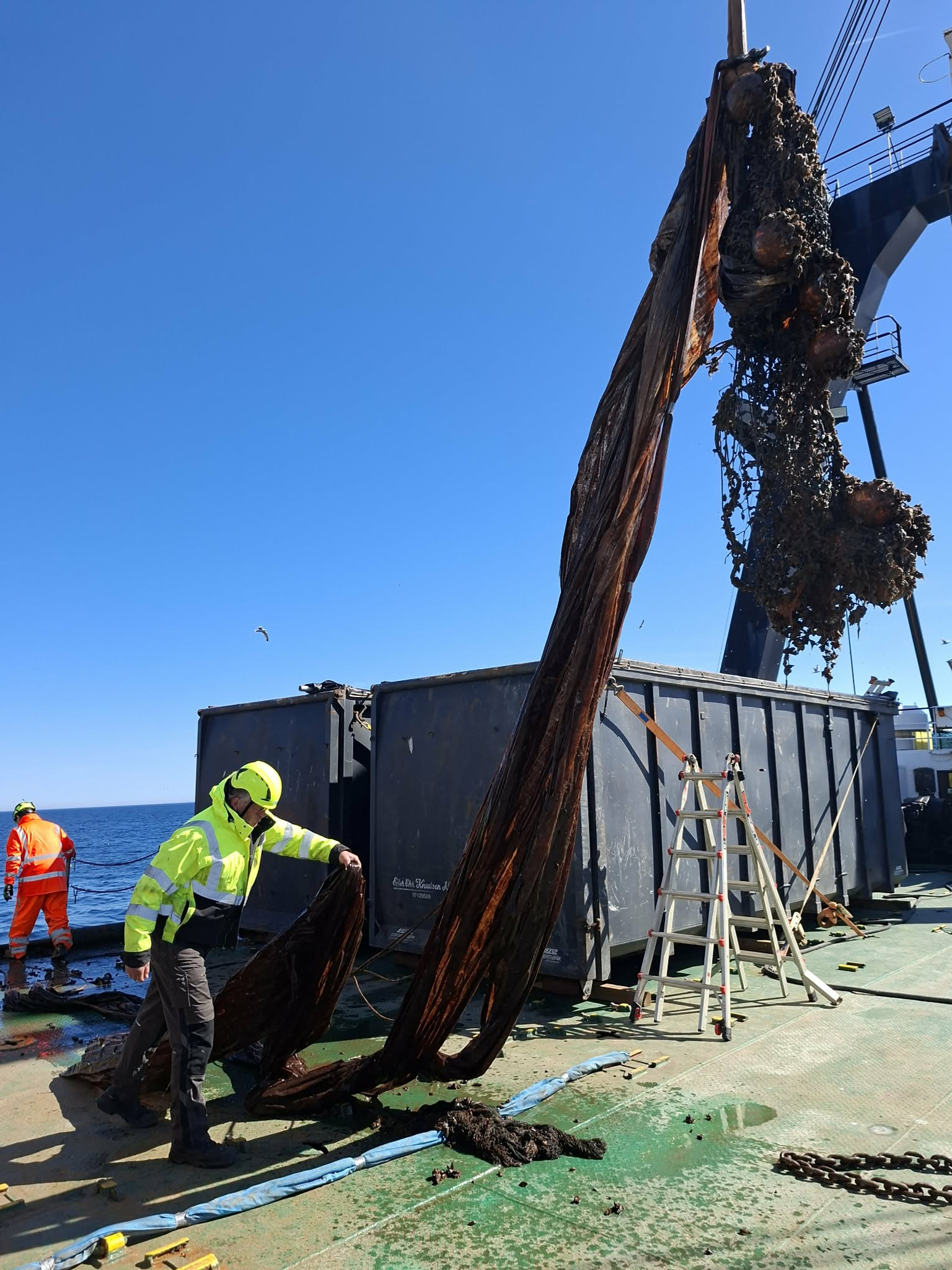Lyftkran drar upp trålnät ur havet och lägger på fartyg. Två personer står på däck.