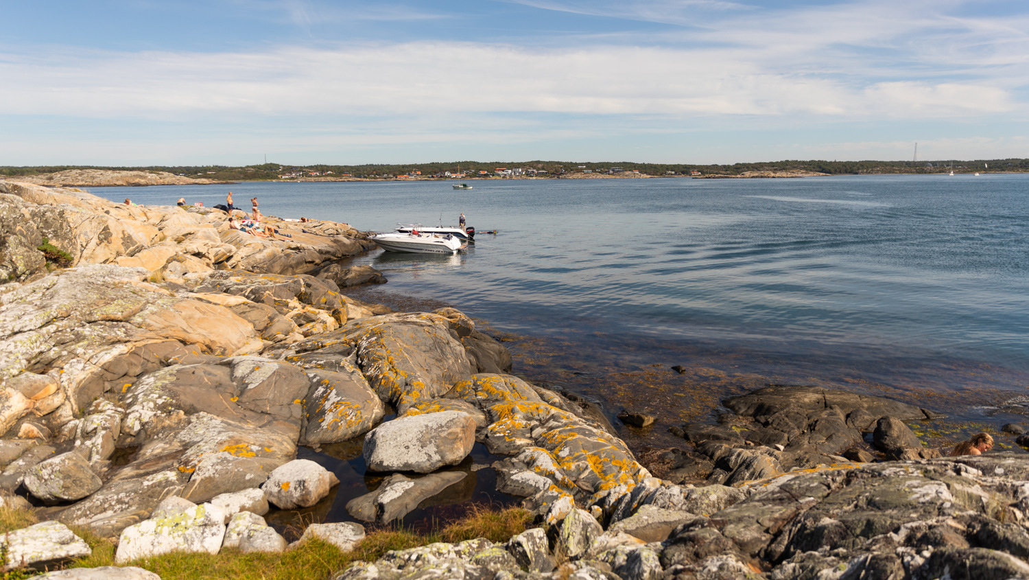 Badgäster på klippor invid hav. I vattenbrynet ligger två motorbåtar förtöjda. I horisonten syns låg, klippig natur med viss växtlighet mot en blå himmel.
