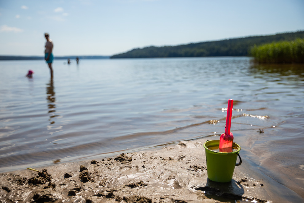 Sandspade och plasthink står på blöt sandstrand framför vatten. I vattnet syns badgäster och i horisonten syns skog mot en blå himmel.