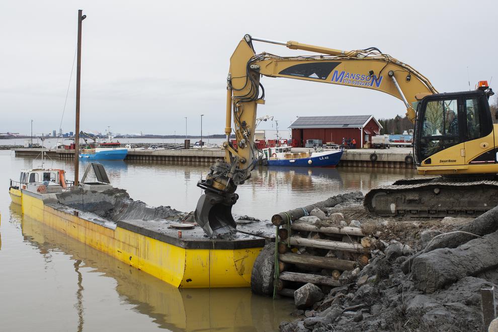 Gul grävmaskin som gräver/muddrar i en båthamn.