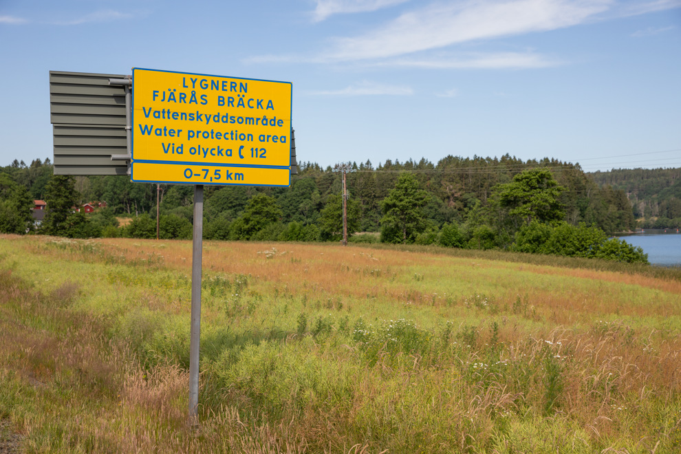 Skylt om vattenskyddsområde invid äng. I bakgrunden syns skog mot en blå himmel med moln.