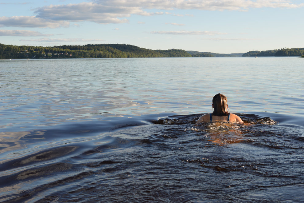 Kvinna simmar i sjö, bort från kameran. I horisonten syns skog.