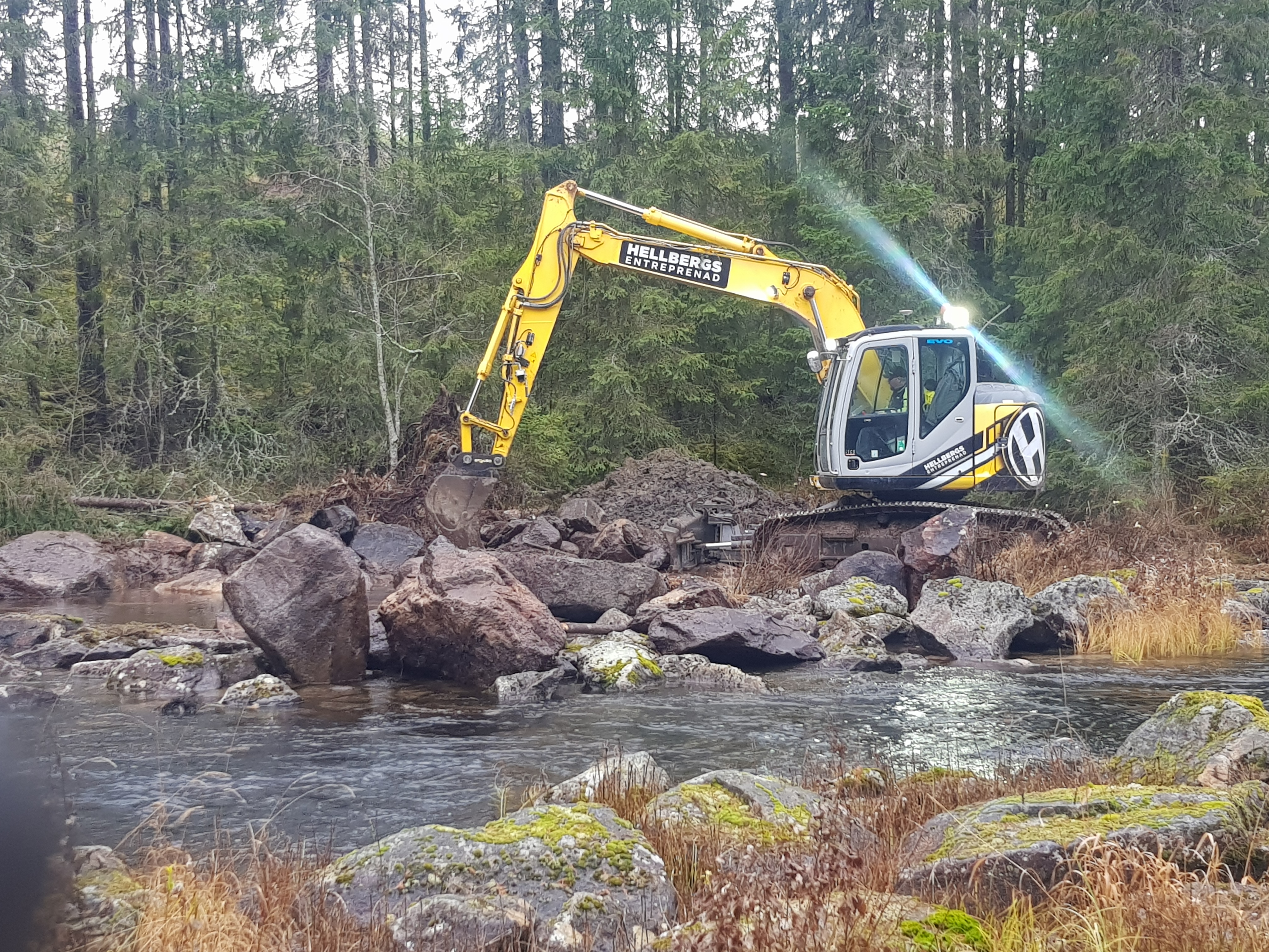 Grävmaskin står på stora klippblock och arbetar i vattendrag, i bakgrunden skog.