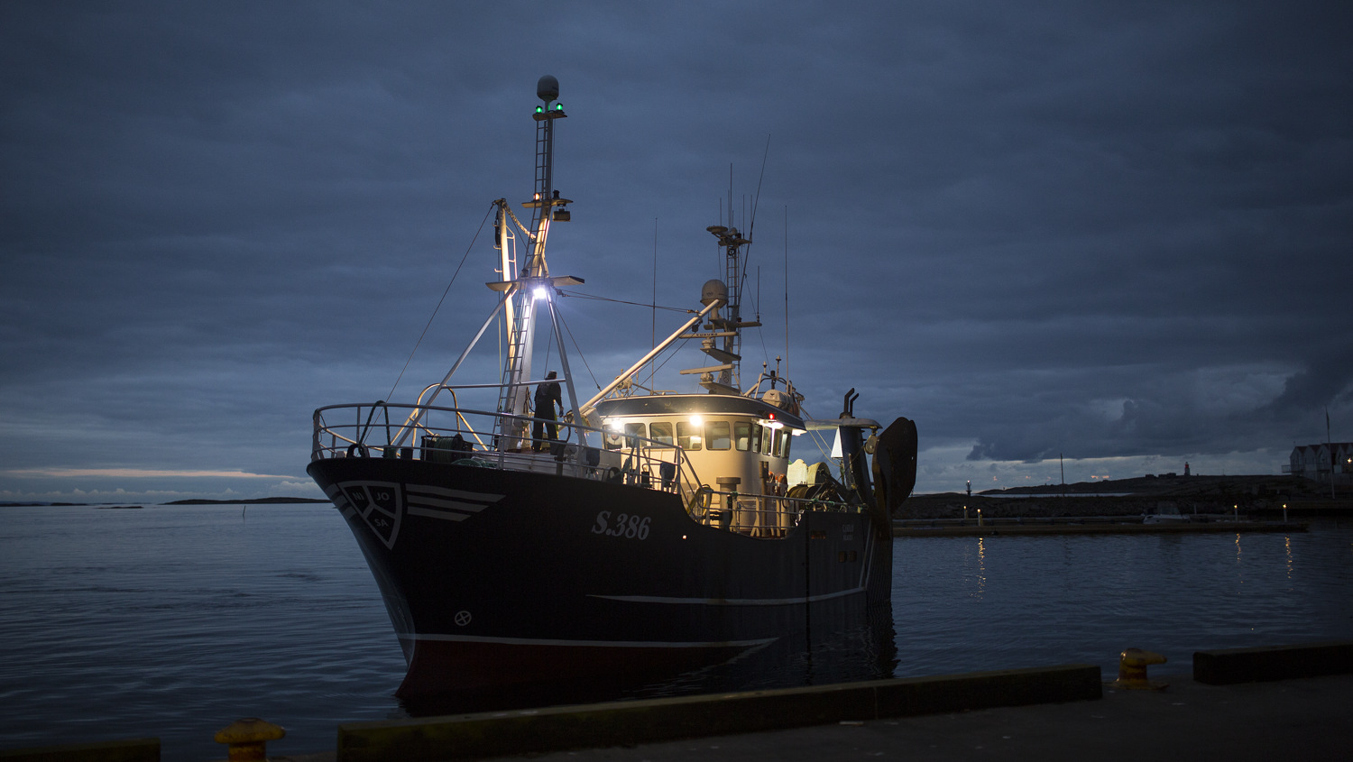 Upplyst fiskebåt i mörker närmar sig kaj