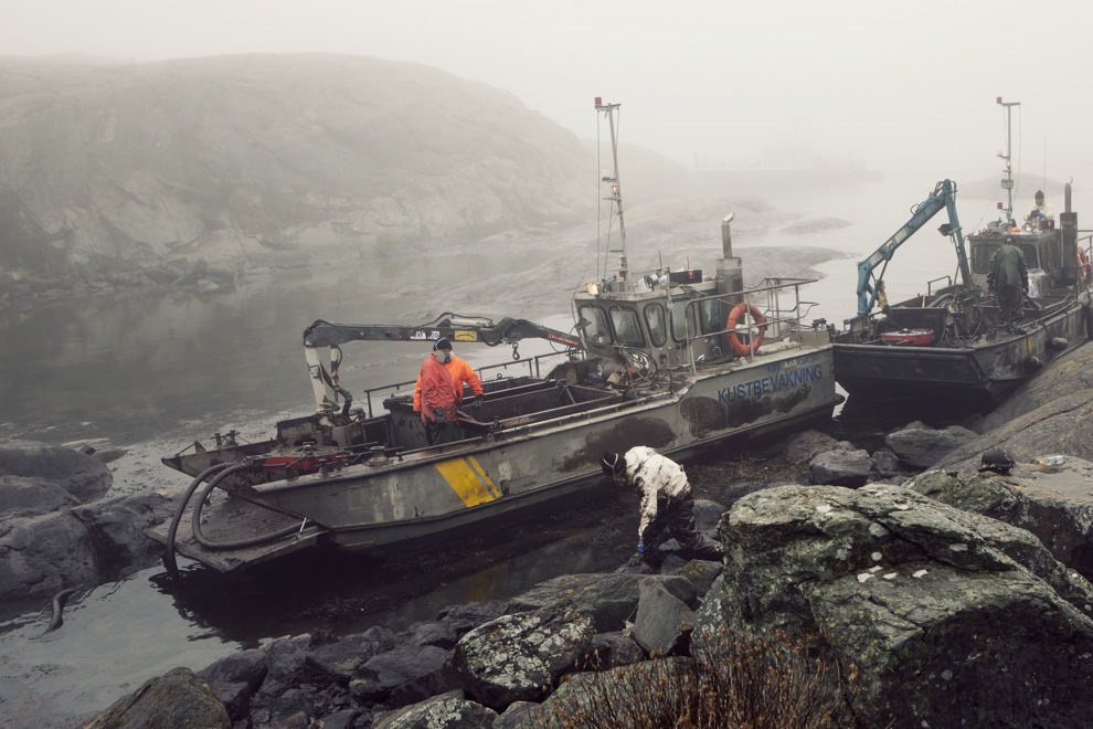 Båtar i vik med mörkt vatten. Klipporna är färgade svarta av olja.