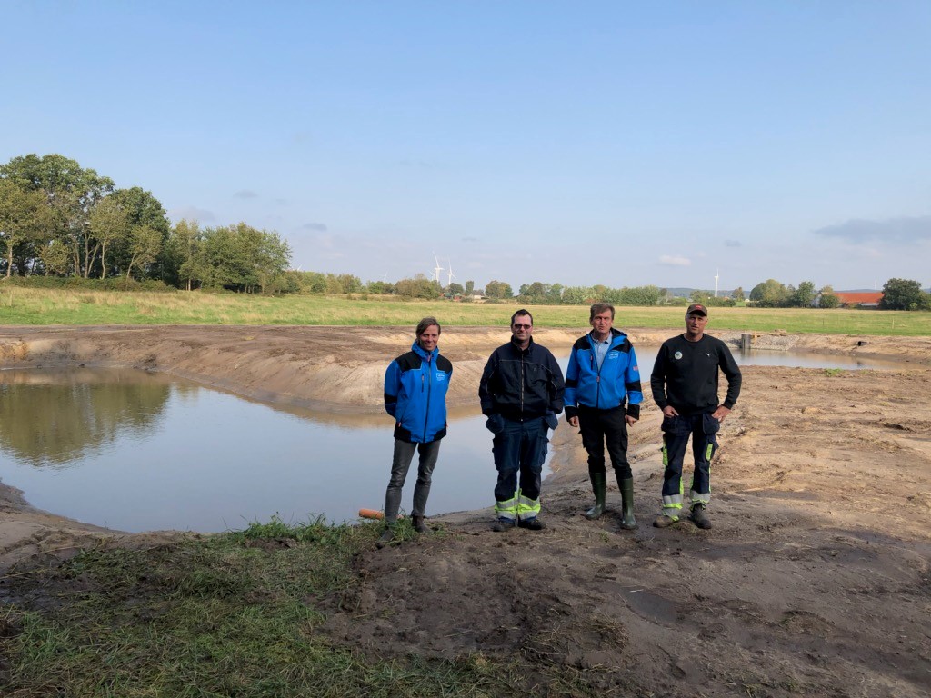 Fyra personer står framför en damm. På marken runt dammen syns ingen växtlighet. 