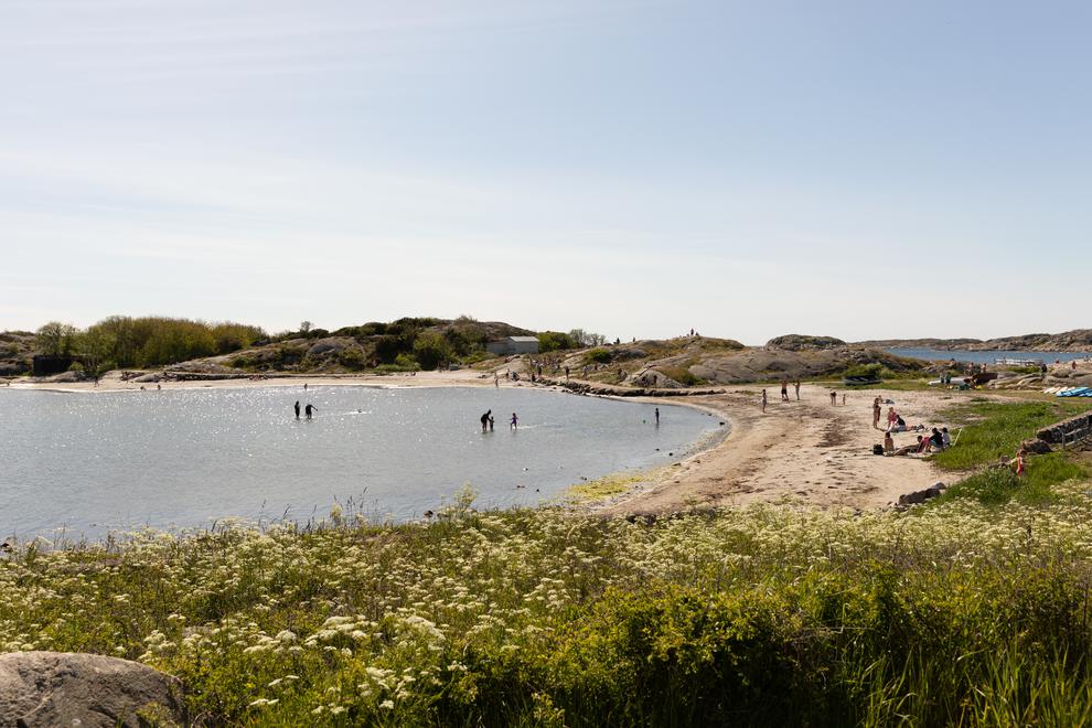 En vik som omges av sandstrand och klippor. I vattnet och på stranden syns badgäster. Landskapet har låg grönskande växtlighet.