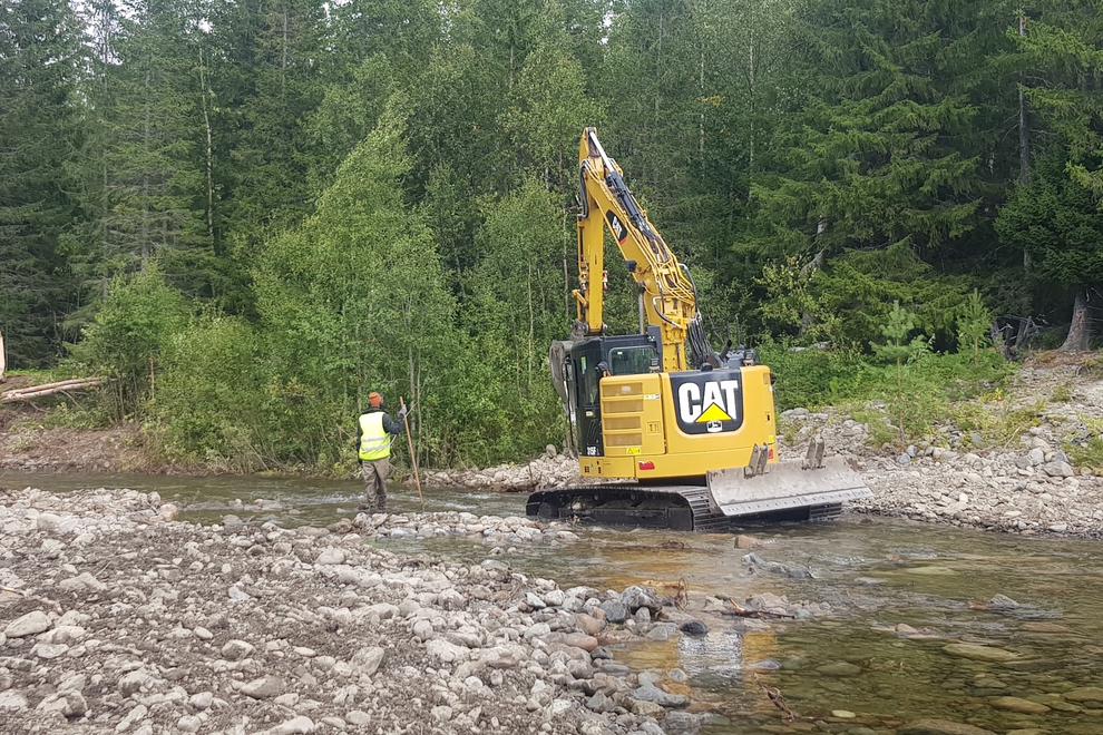 Grävskopa står i vattendrag. Person i varselväst står bredvid. I bakgrund syns barrskog-.